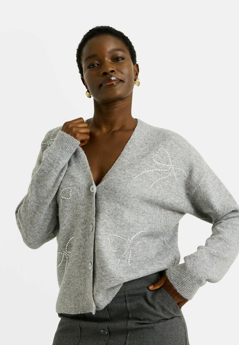 Cardigan en tricot gris avec un col en V, fermeture à boutons et motifs de nœuds en strass décoratifs sur les côtés. Texture douce et coupe décontractée.