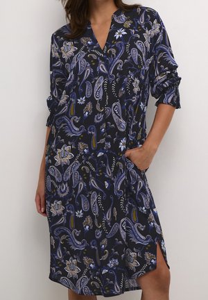 Woman in a dark blue paisley floral dress with long sleeves rolled up, one hand in pocket, standing against a plain light background.