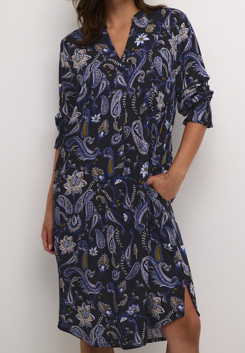 Femme en robe fleurie paisley bleu foncé à manches longues retroussées, une main dans la poche, debout devant un fond clair uni.