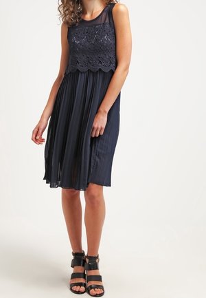 Femme portant une robe noire sans manches avec un corsage en dentelle florale et une jupe plissée, associée à des sandales à talons noires à brides.