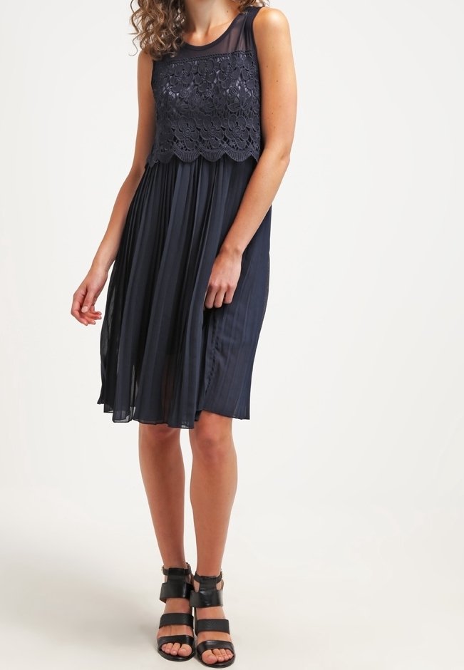 Femme portant une robe noire sans manches avec un corsage en dentelle florale et une jupe plissée, associée à des sandales à talons noires à brides.