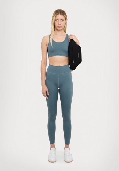 Athletic set in teal: cropped tank with logo band and high-waisted leggings, paired with a black bag. White sneakers complete the look.