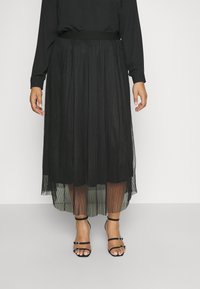 Svart plissert skjørt med en tettsittende midjebånd, semi-transparent lagdelt stoff, kombinert med sorte sandaler med stropper, som avdekker tær og ankel detaljer.
