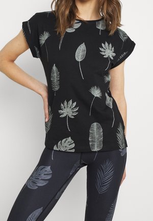 Kvinde iført sort t-shirt og leggings med lysegrå tropiske bladmønstre, poserer med den ene hånd på hoften mod hvid baggrund.