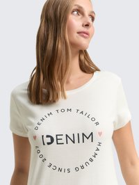 Camiseta blanca de manga corta con un gráfico redondo que tiene "DENIM" en texto negro en negrita y "DENIM TOM TAILOR HAMBURG DESDE 2007" en un diseño circular.