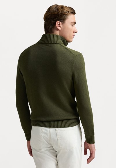 Mørkegrøn strikket turtleneck sweater med ribbet krave, manchetter og kant, båret med lyse bukser.