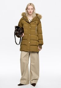 Chaqueta acolchada verde oliva con capucha forrada de piel, con bolsillos con cremallera y botones de presión. Combinada con pantalones anchos beige y un bolso marrón.