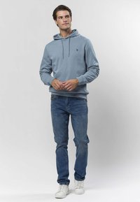 Svetlo modra jopa s kapuco in nastavkom za zategovanje, sprednjo nalepko in rebrastimi manšetami, kombinirana z modrimi kavbojkami in belimi supergami. Gladek teksturni tkanine.