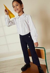 Bluză albă cu mânecă lungă, cu guler volan, asortată cu pantaloni evazați de culoare bleu marin cu un accent floral. Având în mână un zmeu din hârtie galben.