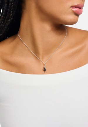 Silberne Halskette mit feiner Kette, die einen detaillierten, verschlungenen Anhänger mit einem kleinen Charm zeigt, vor einem weißen Oberteil.