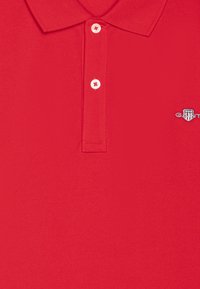 Röd polo-tröja i texturerad tyg med krage, försedd med tre vita knappar och en liten broderad logotyp på bröstet.