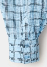 Manche de chemise bleu clair avec motif à carreaux noir et blanc, montrant un poignet boutonné avec deux boutons bleus.