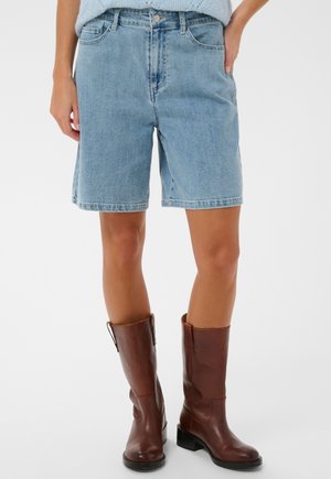 Femeie purtând pantaloni scurți de blugi bleu până la genunchi și cizme maro din piele până la jumătatea gambei, pe un fundal alb.