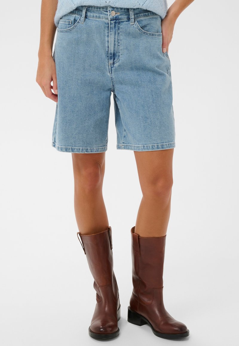 Vrouw die lichtblauwe denim korte broek tot aan de knieën draagt en bruine leren laarzen tot halverwege de kuit, op een witte achtergrond.