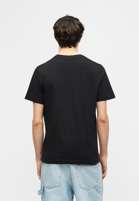 Άνδρας με κοντά σκουρόχρωμα μαλλιά, που φοράει απλό μαύρο κοντομάνικο μπλουζάκι και ανοιχτό μπλε τζιν, στέκεται στραμμένος προς τα πίσω πάνω σε λευκό φόντο.