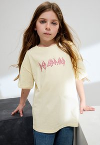 Hellgelbes Baumwoll-T-Shirt mit kurzen Ärmeln, versehen mit pinkfarbenem "DEF LEPPARD"-Schriftzug in einer markanten, kantigen Schriftart. Kombiniert mit blauen Jeans.