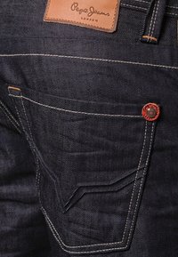 Mörka denimjeans med orange sömmar, en läderflik med varumärkeslogo och en distinkt böjd fickdesign. Har en röd knapp vid midjan.