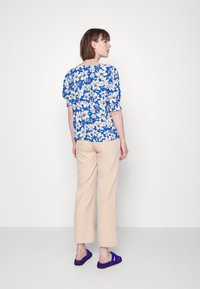 Blusa de padrão floral em azul e branco, de corte solto com mangas bufantes. Calças beijas de perna reta e sandálias roxas de slide completam o look.