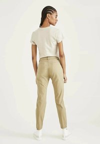 Pantaloni beige aderenti con un taglio classico, abbinati a una t-shirt grigia chiara, impreziositi da tasche posteriori discreti e sneakers bianche. Texture liscia.