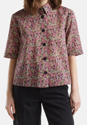 Kvinde iført en kortærmet skjorte med knapper, der har et tæt mønster af pink, gule og hvide blomster, kombineret med sorte bukser.