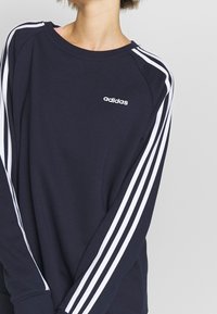 Sudadera azul marino con diseño de tres rayas blancas a lo largo de las mangas, cuello redondo y logotipo bordado de Adidas en el pecho. Textura de tela suave.