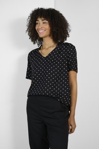 Schwarze Bluse mit Punkten, V-Ausschnitt, kurzen Ärmeln und lockerer Passform, aus leichtem Stoff, kombiniert mit schwarzen Hosen.
