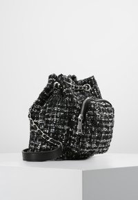 Sac en tweed noir et blanc avec une finition texturée, doté d'une bandoulière en chaîne, d'une poche zippée et d'une fermeture à cordon en haut.