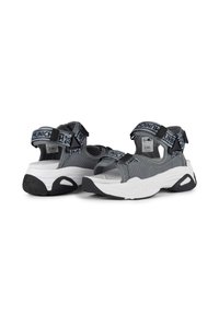Sandales en tissu gris avec des brides ajustables arborant un logo, semelle intermédiaire blanche et semelle extérieure en caoutchouc noir. Détails texturés et matériau respirant.