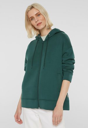 Young woman with short blonde hair wearing a dark green zip-up hoodie and white pants against a plain light background.