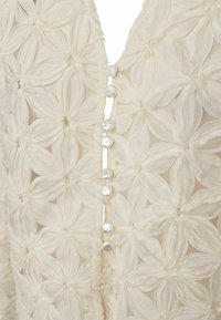 Krems krāsas ziedu mežģīņu apģērbs ar dziļu V-veida kaklu, rotāts ar nevienmērīgiem pērlīšu pogām gar priekšējo plati.