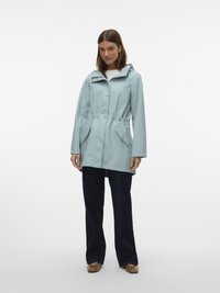 Nainen pukeutuneena vaaleansiniseen hupulliseen sadeviittaan, mustiin leveälahkeisiin housuihin ja ruskeisiin kenkiin, seisoo yksinkertaisen valkoisen taustan edessä.