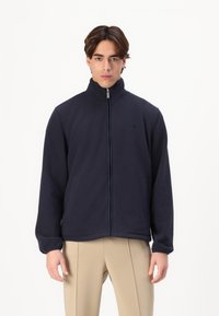 Jachetă din polar bleumarin cu guler înalt și fermoar complet, dotată cu buzunare laterale și un branding subtil pe piept. Textură moale și croială lejeră.