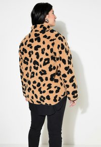 Kabát s leopardím potiskem vyrobený z hnědého fleece materiálu s černými skvrnami, zaobleným límcem a kontrastním černým lemem. Oblečený přes černé kalhoty.