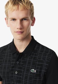 Czarna koszulka polo z geometrycznym wzorem w odcieniach szarości, z zielonym wyszywanym logo krokodyla na piersi oraz klasycznym kołnierzykiem.