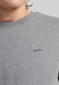Szara bluza z miękkiego, teksturowanego materiału, z prążkowanym dekoltem i małym, niebieskim haftowanym logo na piersi.