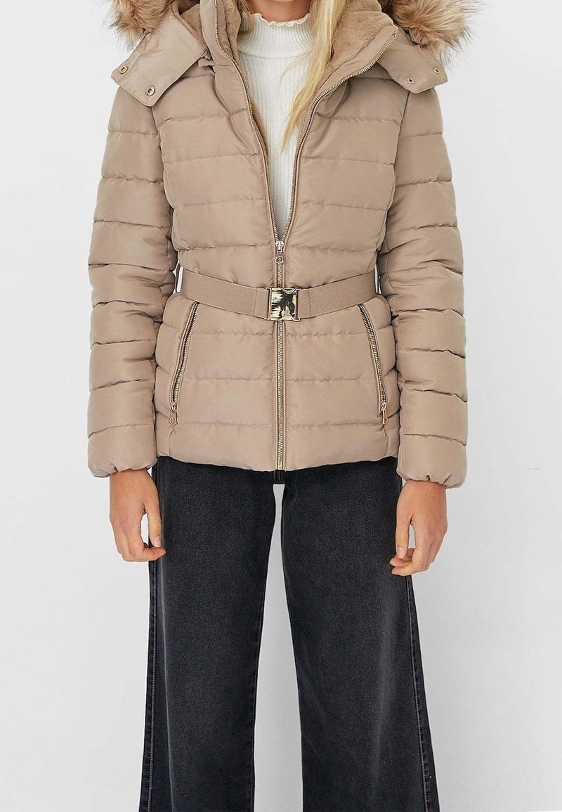 Lichtbeige gewatteerd jack met een capuchon met bontvoering, ritssluiting, tailleband met ceintuur en horizontale stiksels. Draagt het over een wit shirt en donkere spijkerbroek.