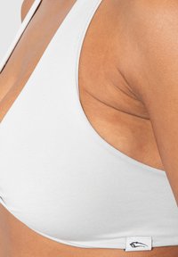 Vit triangulär bralette med tunna band, tillverkad av ett mjukt tyg. Har en logotag vid nederkanten. Mjuk textur och enkel design.