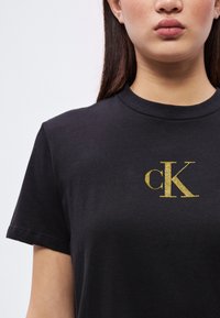 Jeune personne portant un t-shirt noir avec un logo "cK" doré et scintillant centré sur la poitrine, sur un fond blanc uni.