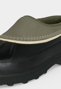 Gros plan d’une chaussure noire et vert olive imperméable à enfiler, avec une semelle en caoutchouc épaisse et des détails de couture blancs.
