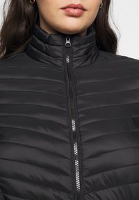 Giacca nera trapuntata con collo alto, dotata di una cerniera frontale e una texture liscia e lucida. Cuciture orizzontali e design minimalista.