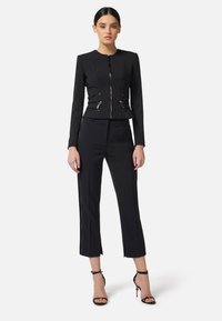 Traje negro a medida que cuenta con una chaqueta ajustada con cierre de cremallera y bolsillos con cremallera, combinada con pantalones recortados. Acentuado con detalles en dorado.