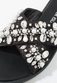 Svart sandal med en rem prydd med juveler, inklusive klara och iriserande kristaller, pärlor och ett övre lager av texturerat tyg. Platt sula.