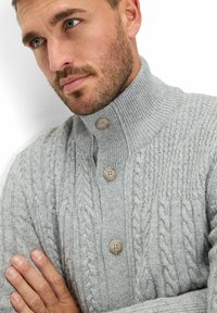 Hellgrauer Kabelstrickpullover mit hohem Kragen, ausgestattet mit drei großen Knopfverschlüssen und strukturierten Mustern im Gewebe.