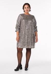 Abito scintillante con paillettes nei toni argento e bronzo, di lunghezza al ginocchio e con maniche lunghe. Indossato con tacchi in vernice nera e collant neri trasparenti.