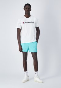 Maglietta bianca con logo "Champion", abbinata a pantaloni corti azzurri chiari. Accessori includono sneakers alte bianche e calzini bianchi con logo.