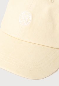 Ανοιχτό κίτρινο καπέλο μπέιζμπολ με καμπυλωτό γείσο και λευκό κεντημένο κυκλικό έμβλημα που παρουσιάζει σχέδιο τετράφυλλου λουλουδιού.