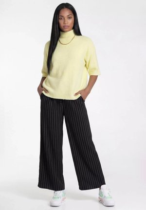 Kobieta stoi z rękami w kieszeniach, ubrana w jasnożółty golf, czarne szerokie spodnie w cienkie paski, białe trampki i złoty łańcuszek na szyi.