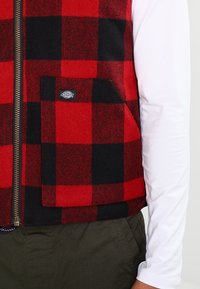 Röd och svart rutigt zip-up väst med ficka fram och Dickies-logotyp, bärs över en vit långärmad skjorta och mörkgröna byxor.