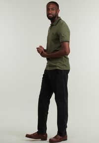 Camisa polo verde oliva con acentos de color negro en el cuello, combinada con pantalones negros y zapatos marrones. El modelo está en una posición relajada.