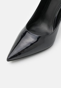 Chaussure à talon haut vernie noire à bout pointu avec une surface en cuir lisse et des coutures visibles le long du bord intérieur.